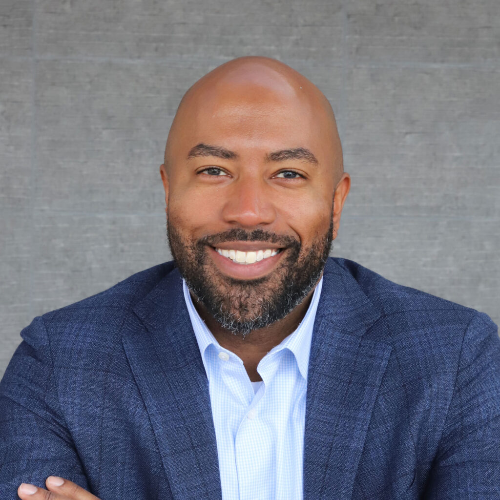 Sean Perryman, smiling in front of a grey backgdrop wearing a dark blue suit jacket and light blue collared shirt.