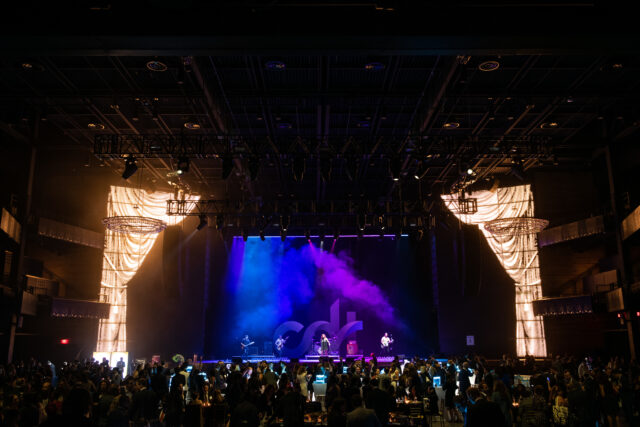Tech Prom guests interact throughout the event, while the stage is awash in music from the band and bright purple and blue lights.