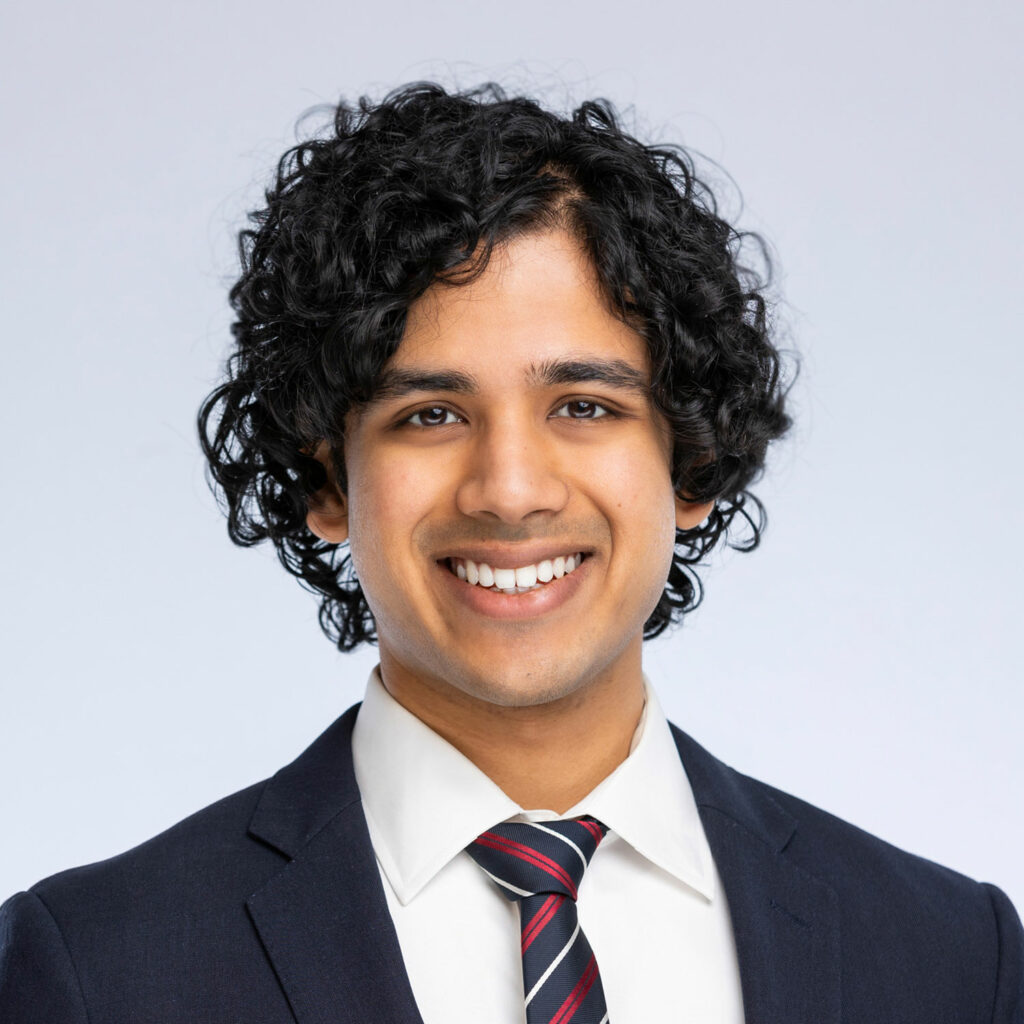 Chinmay Deshpande, smiling wearing a dark suit jacket and red, white and blue striped tie.