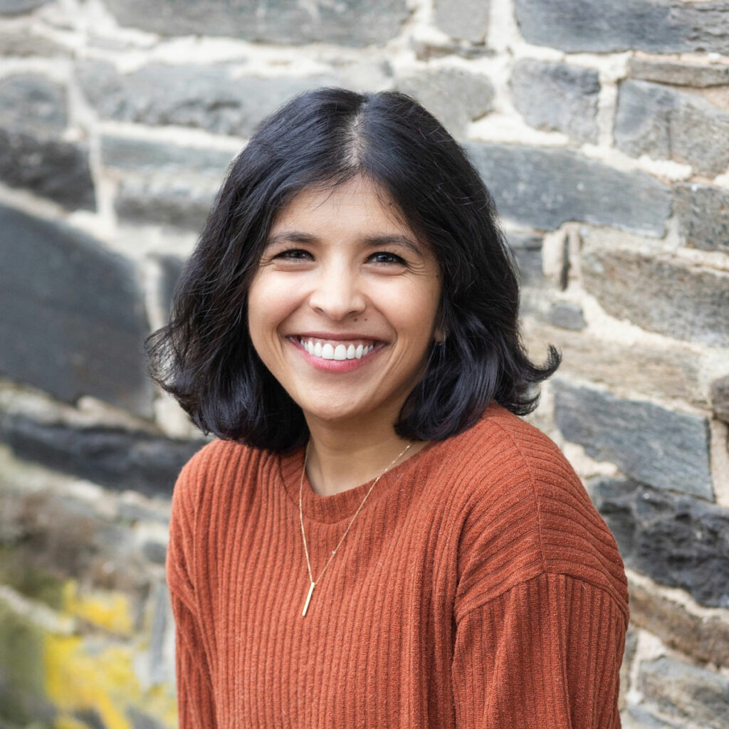 Chinmayi Sharma, smiling wearing a burnt orange top and necklace in front of a grey stone background.