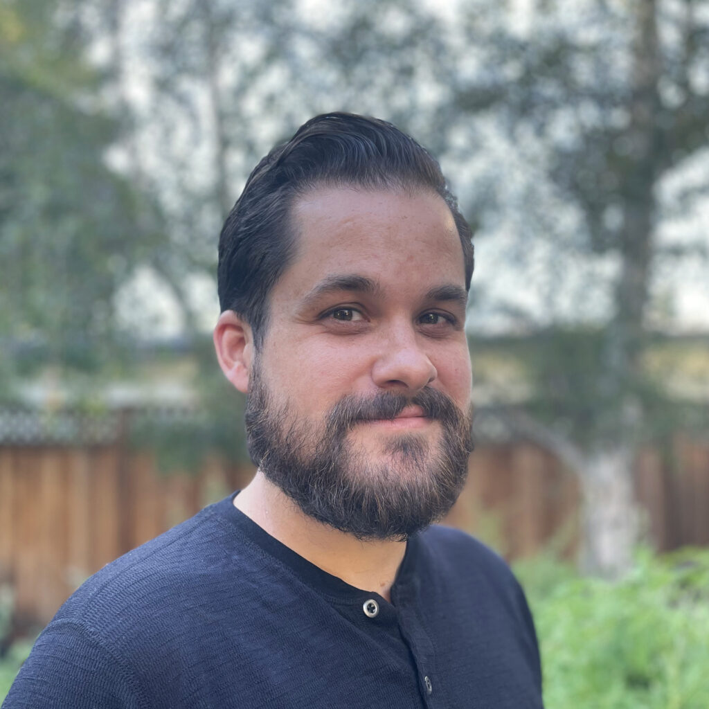 Dave Willner, wearing a blue shirt in front of a green outdoor setting and wooden fence.