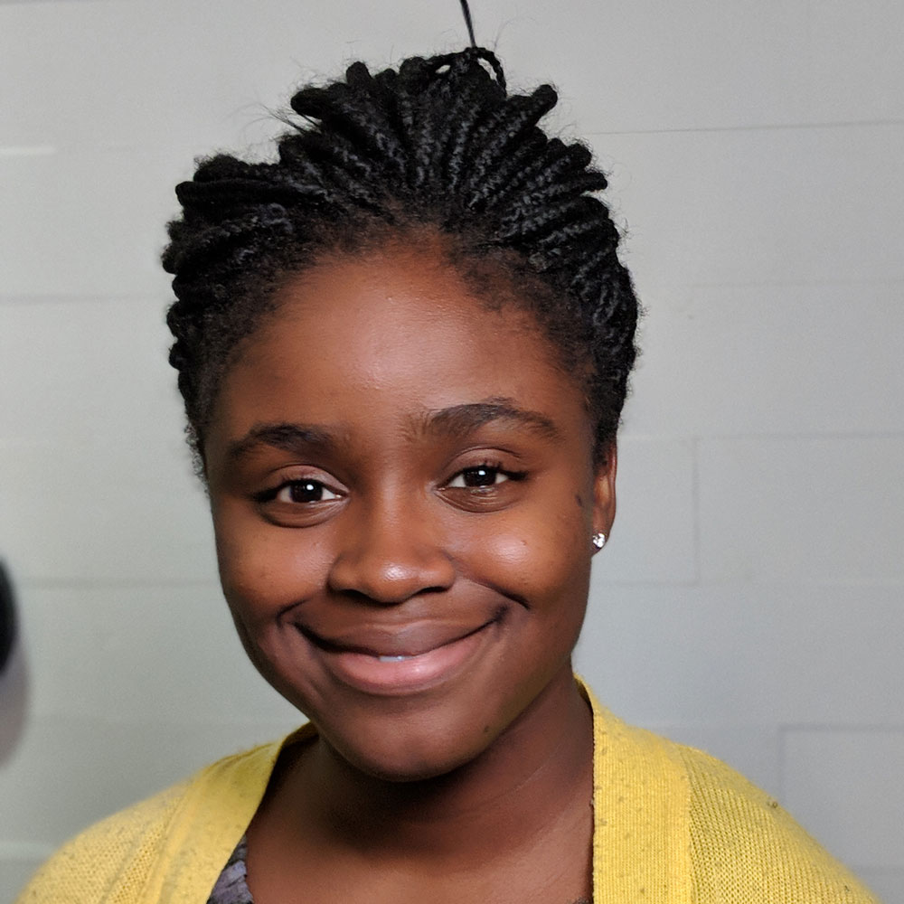 Deb Raji, wearing a yellow top smiling in front of a grey background.