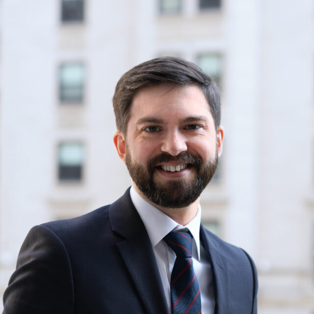 Tim Harper, smiling with a blue jacket, white shirt and blue tie, in front of city building background.