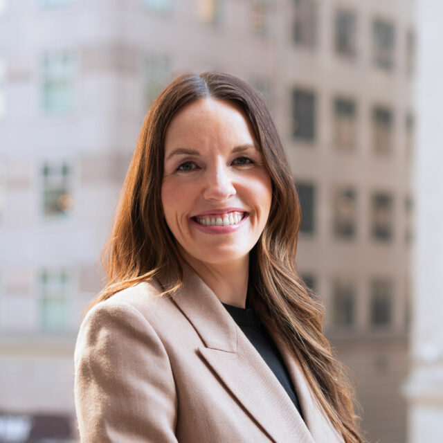 Elizabeth Laird, smiling wearing a light brown jacket and black top, outdoors in front of a city background.