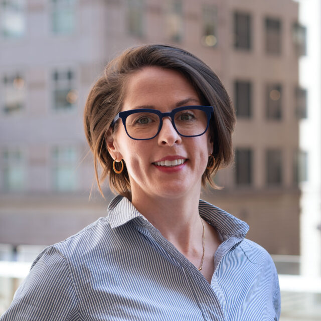 Amy Winecoff, smiling in dark blue rimmed glasses, short hair and a striped collared shirt outside in a sunny cityscape.