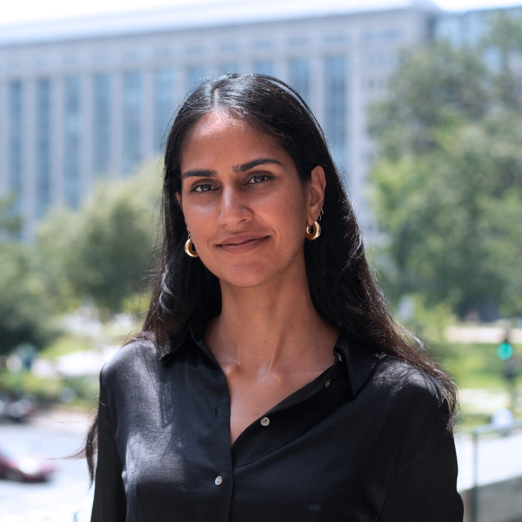 Aliya Bhatia, smiling outdoors wearing a black silk top and gold earrings.
