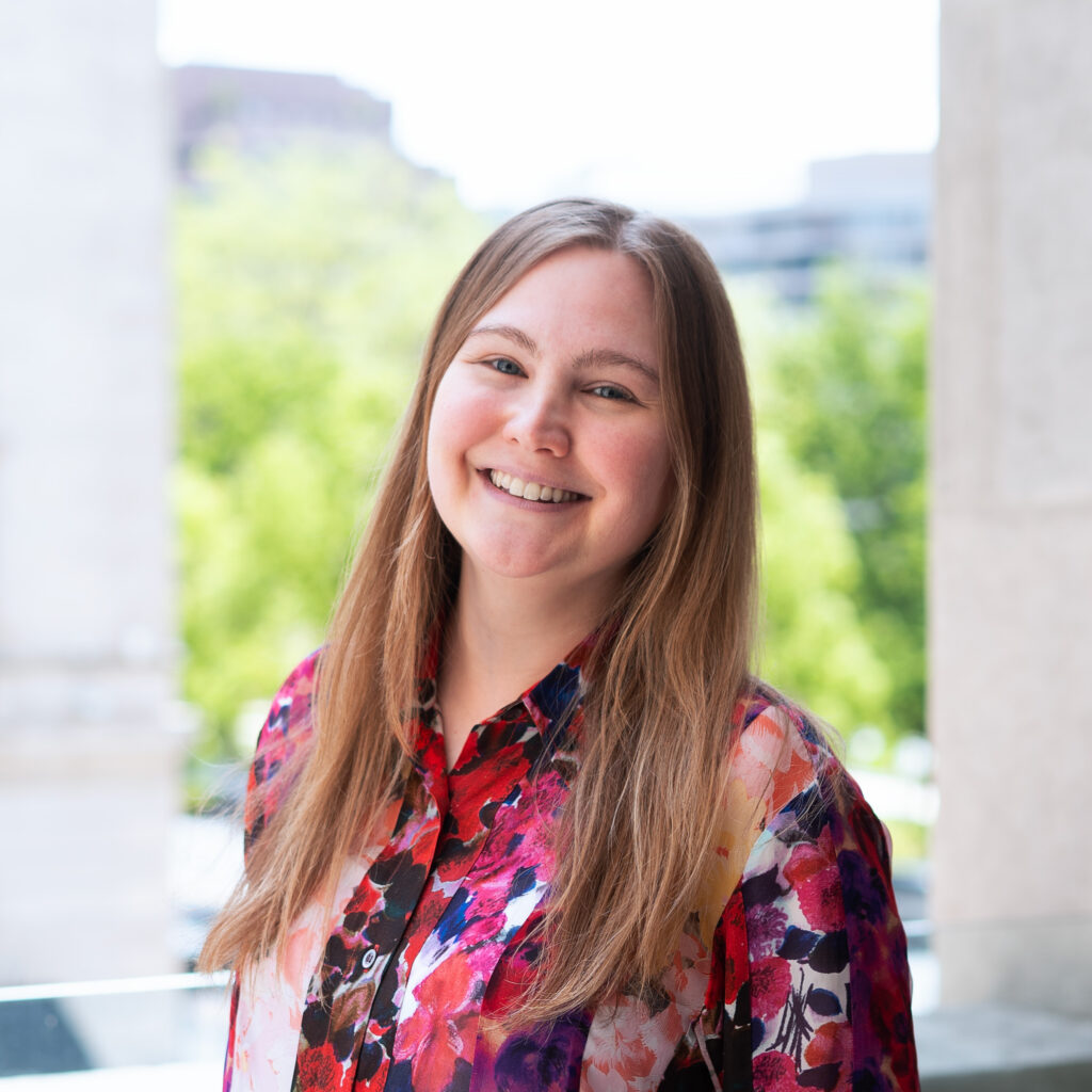 Elizabeth Seeger, smiling in a multi-color top, standing outside in a cityscape with trees behind her.