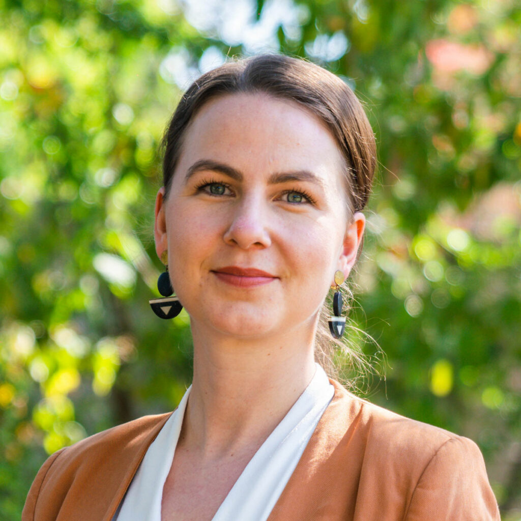 Sabine Witting, wearing a tan jacket and white top in front of a leafy green background.