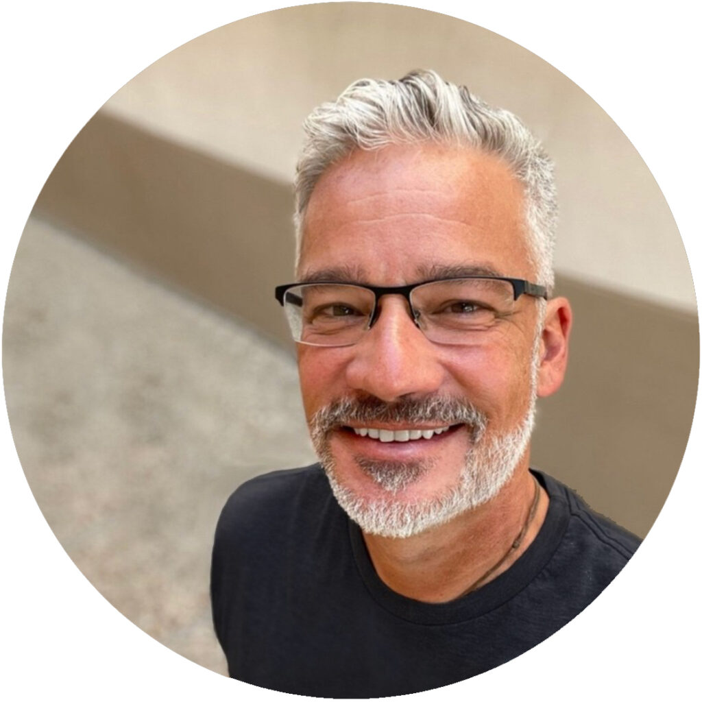 Edward Perez, smiling wearing black rimmed glasses in front of a light colored wall.