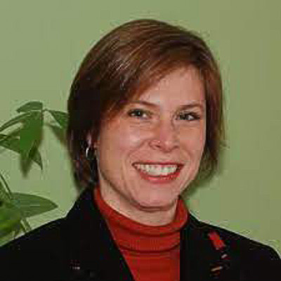 Megan Gray. DuckDuckGo. Wearing a red turtleneck with a dark jacket, in front of a light green background (and a green leafy plant).