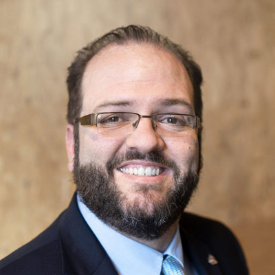 Joe Hall. Internet Society. Wearing a light blue collared shirt and dark suit, in front of a light brown background.
