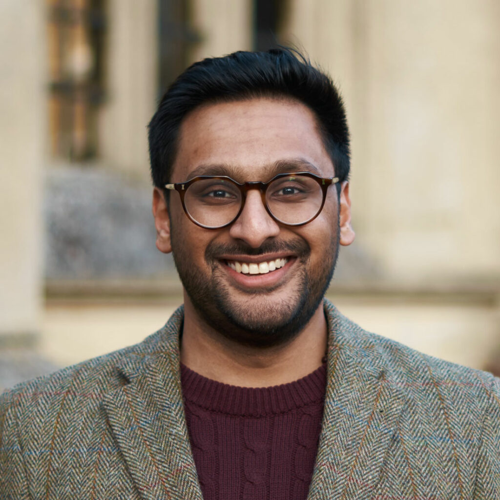 Umang Bhatt, smiling wearing black round glasses and a light colored jacket standing in front of a building facade.
