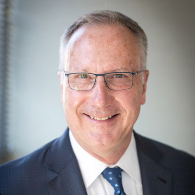 Robert Holleyman. Crowell & Moring. Wearing glasses and a white collared shirt and blue suit, in front of a light colored background.