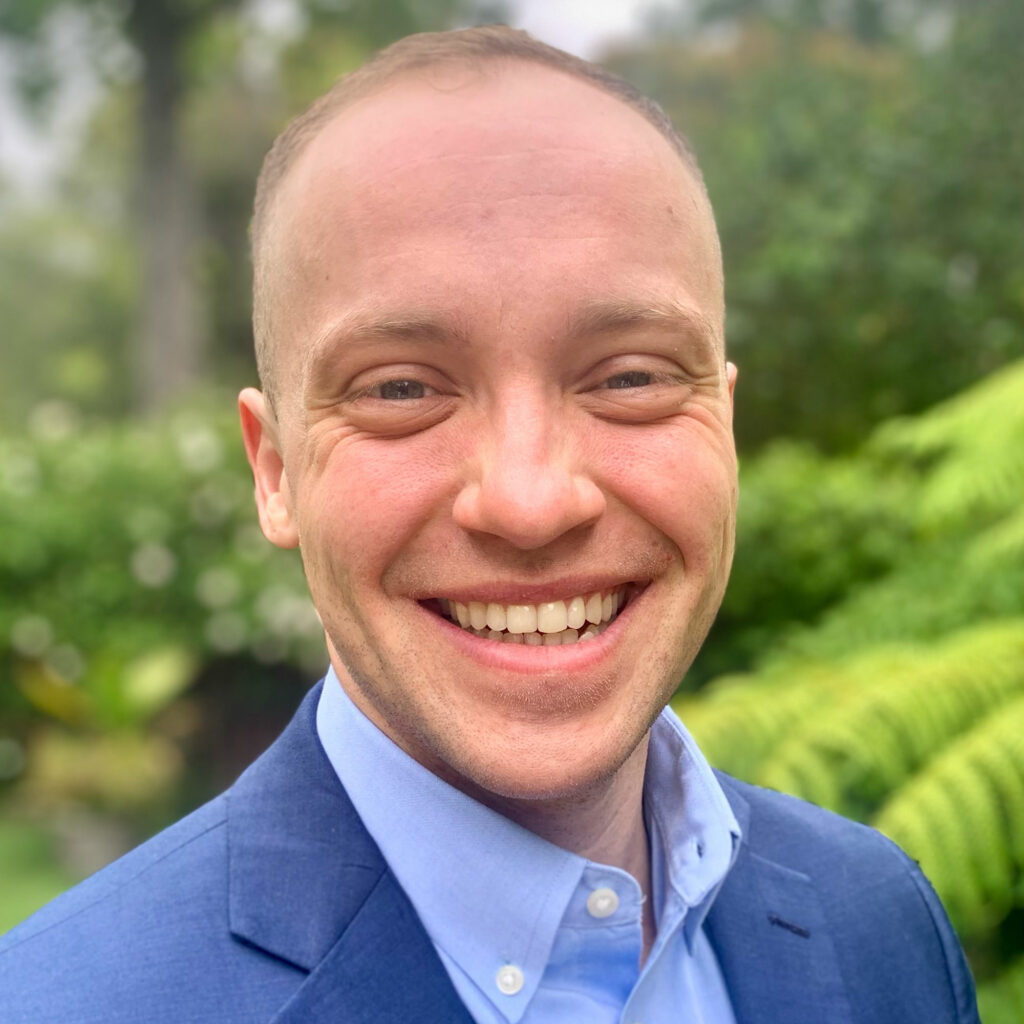 Quinn Anex-Ries, smiling while wearing a blue jacket and collared shirt, outside in a leafy green background.