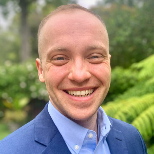 Quinn Anex-Ries, smiling while wearing a blue jacket and collared shirt, outside in a leafy green background.