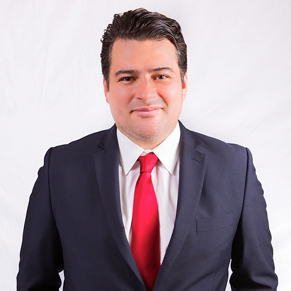 Juan Proaño, in a dark suit and bright red tie, in front of a white backdrop.