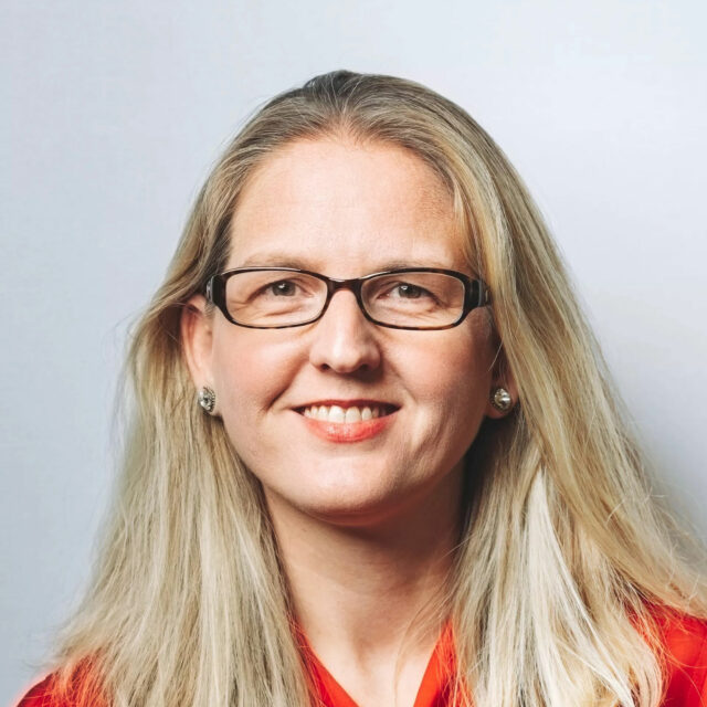 Kate Ruane, wearing glasses and a red top, smiling in front of a white wall