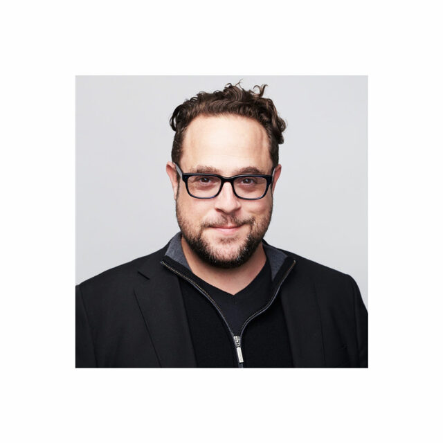Kevin Bankston, wearing black glasses, a jacket and shirt in front of a white background.