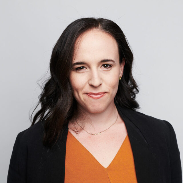 Miranda Bogen, wearing a burnt orange top and black jacket, smiling in front of a grey background.