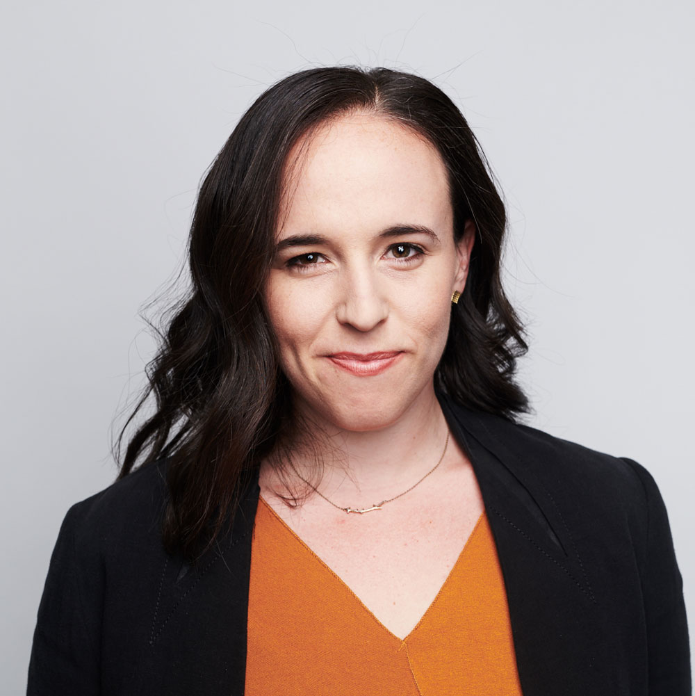 Miranda Bogen, wearing a burnt orange top and black jacket, smiling in front of a grey background.