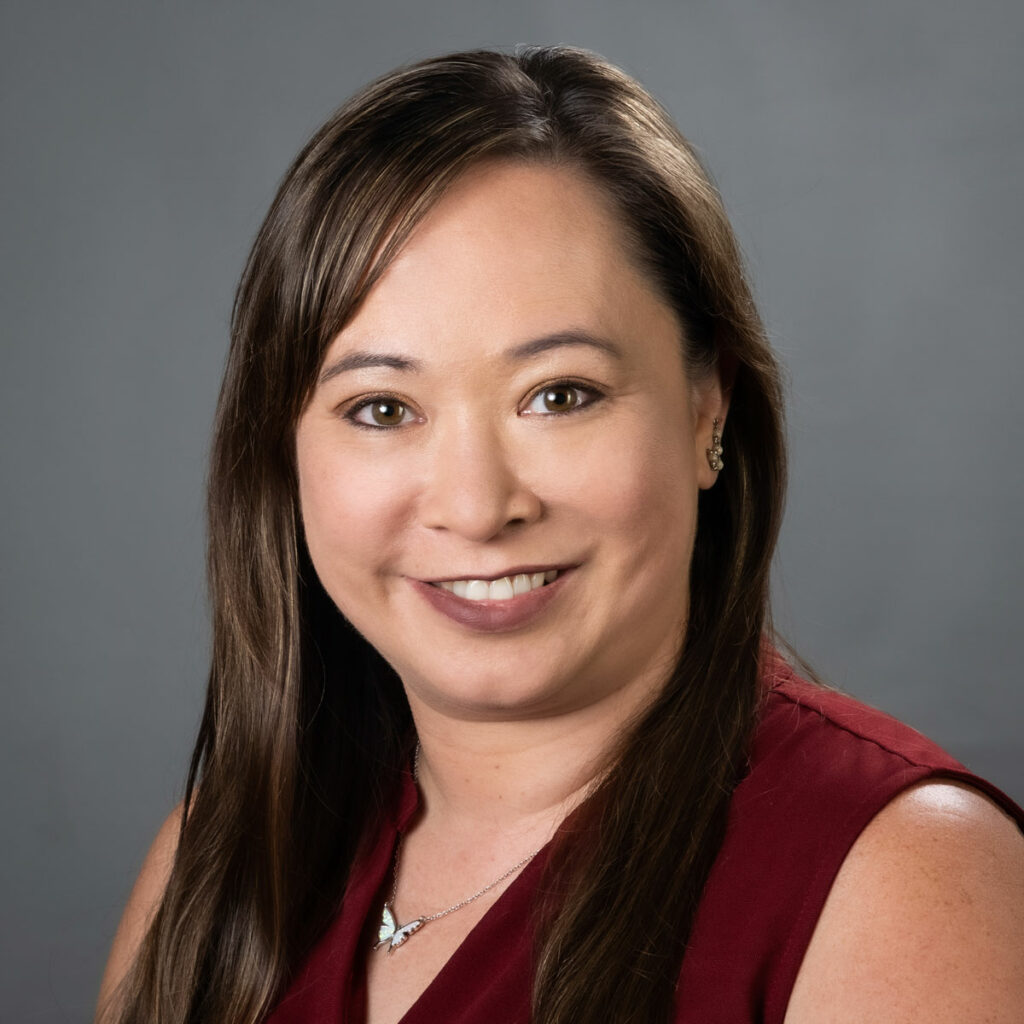 Pamela Wisniewski, smiling wearing a deep red top in front of a dark grey background.
