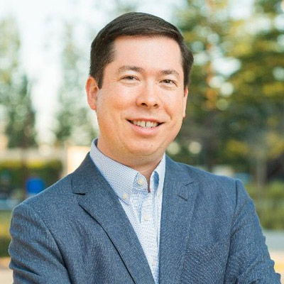 David Quinalty. Waymo. Wearing a light blue collared shirt and a light blue / gray suit, in front of a leafy green background.
