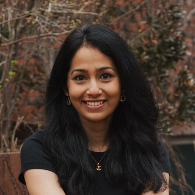 Ruchika Joshi, smiling wearing a black top and necklace in front of a fall outdoor background.
