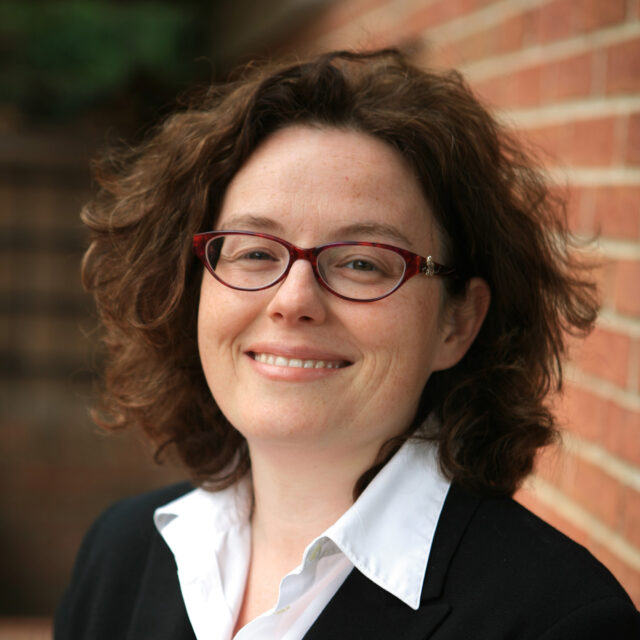 Sarah Zolad, wearing glasses, a white collared shirt and a black sweater, smiling in front of a brick outdoor setting.