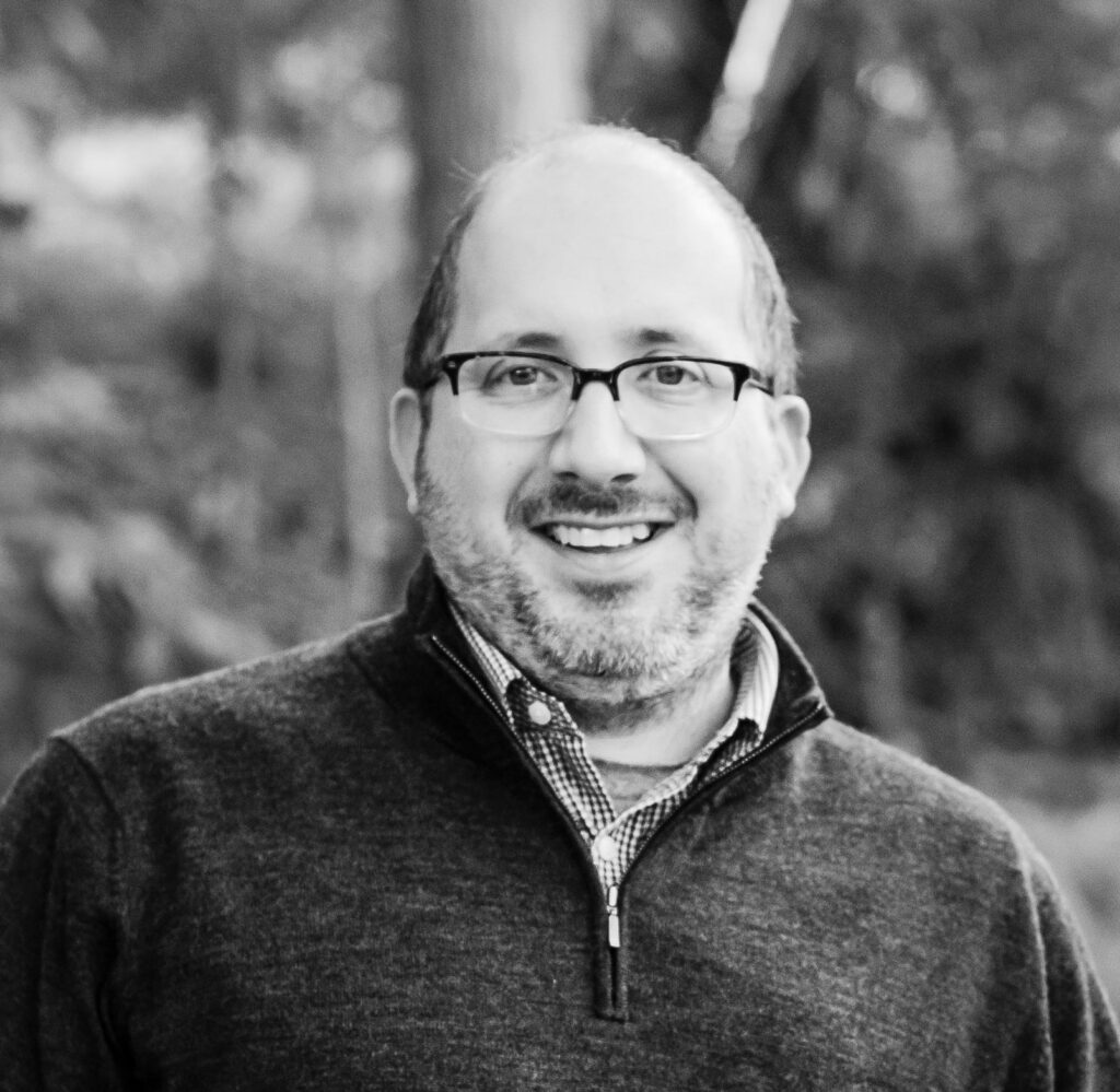 Shawn Bone, smiling in rimmed glasses and a collared shirt and sweater (outdoors, in a black-and-white photo).