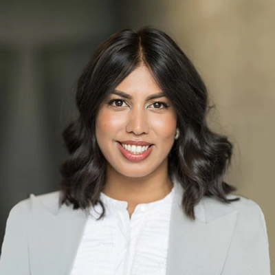 Nabiha Syed. The Markup. Wearing a white collared shirt and a light grey suit, in front of a light brown background.