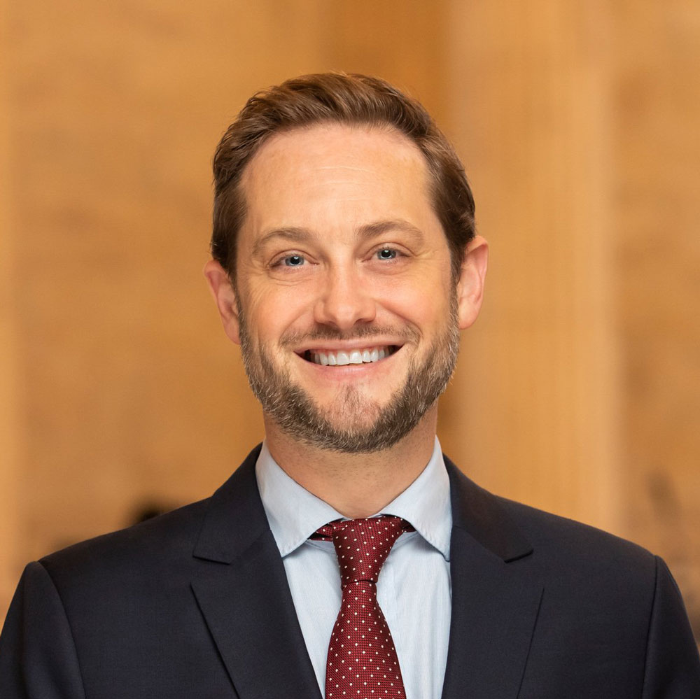 Travis Moore, wearing a dark jacket and dark red tie, smiling in front of an indoors setting.