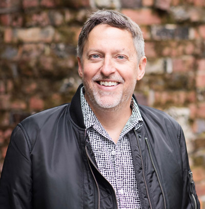Richard Whitt. GLIA Foundation. Wearing a light grey collared shirt and a dark grey jacket, in front of a brown brick background.