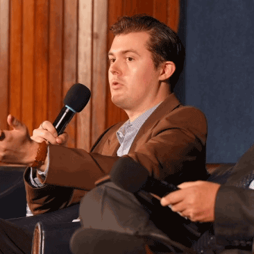 Justin Sherman, in a brown blazer holding a microphone speaking up on a stage.